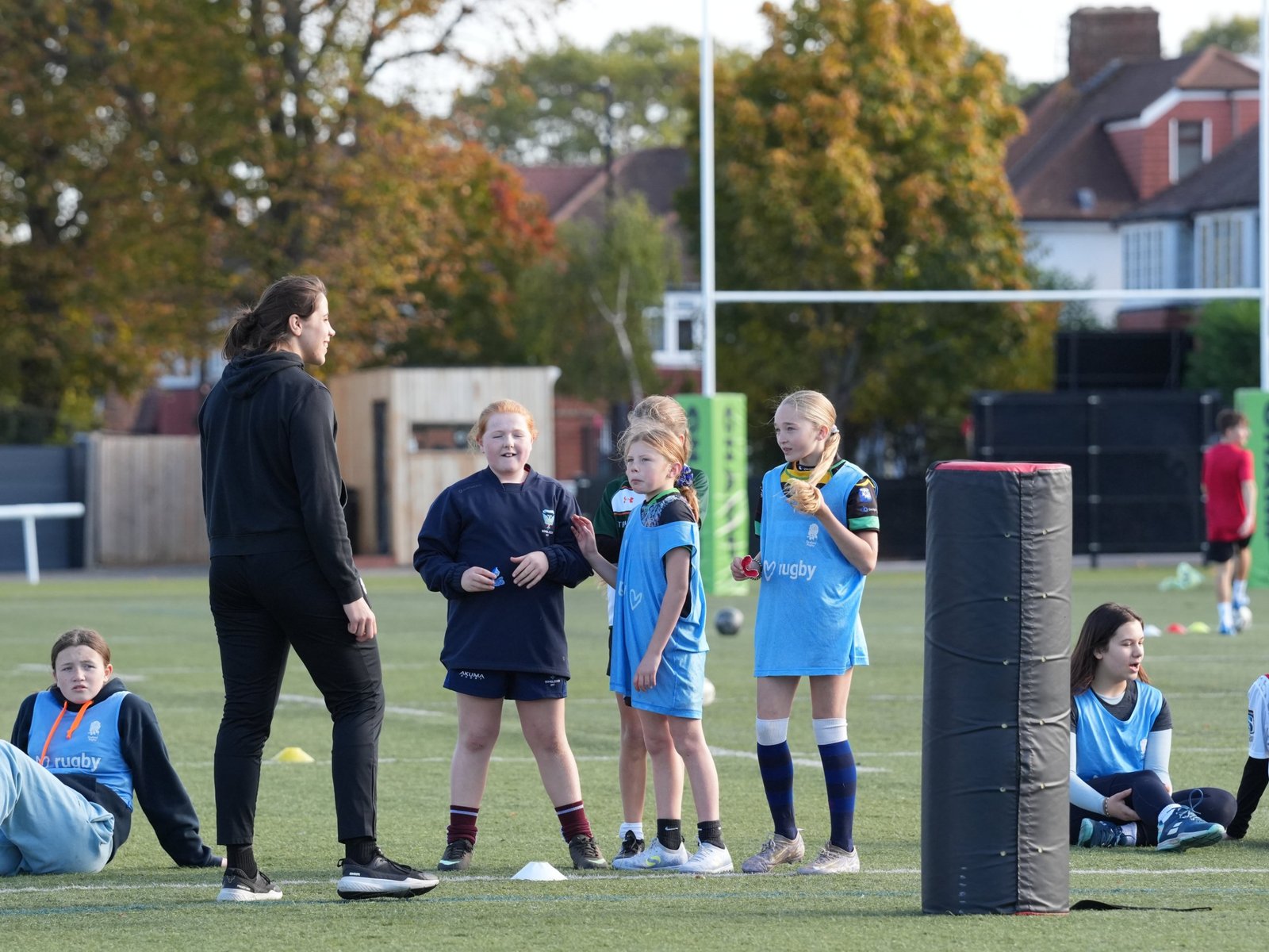 A coach in a black tracksuit stands on an artificial pitch talking to four girls in light blue bibs, with a tackle shield in the foreground and goalposts behind.
