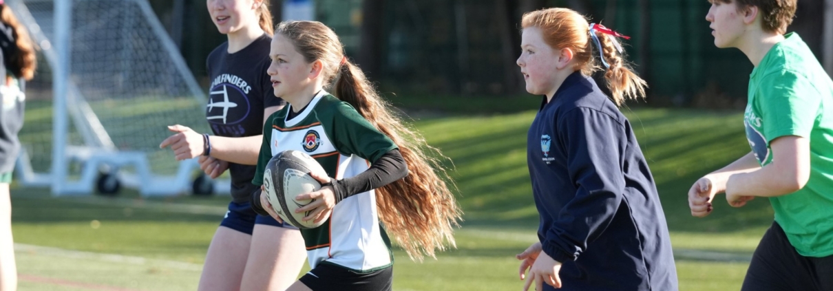 A girl in a green and white rugby shirt runs with the ball, with two girls and a coach following behind on an artificial pitch.