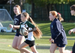 A girl in a green and white rugby shirt runs with the ball, with two girls and a coach following behind on an artificial pitch.