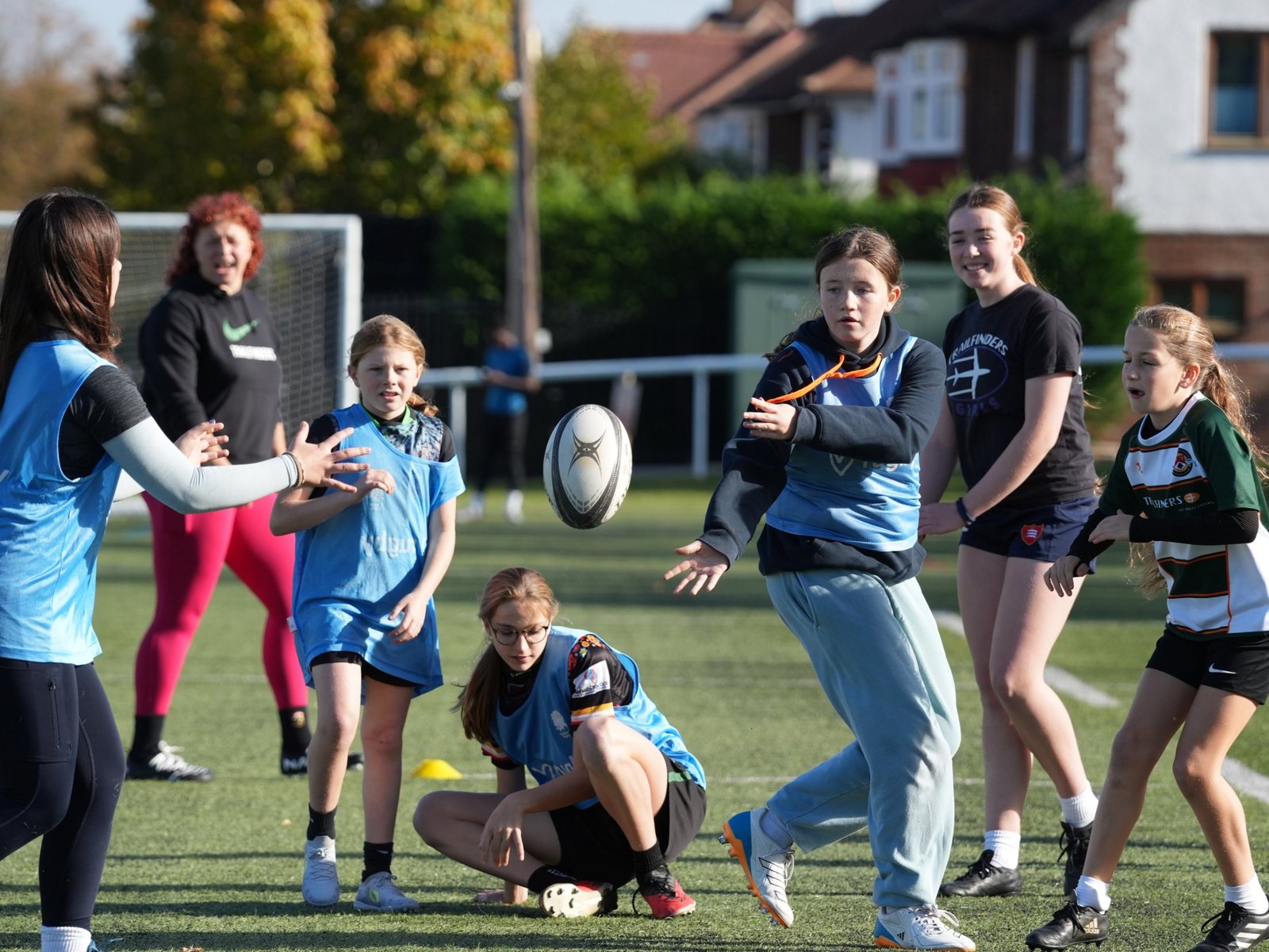 A girl in a light blue bib passes a rugby ball to a girl on the left, while other girls and two coaches in black tops watch on an artificial pitch.